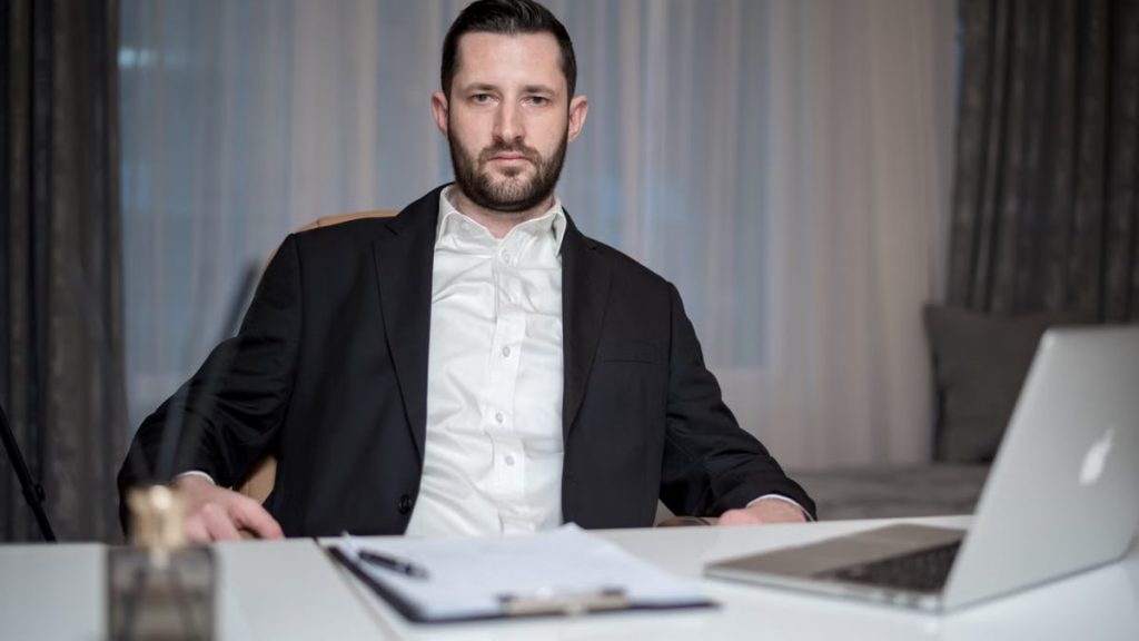 A serious bearded man in a black suit sits behind a desk with a laptop and documents.